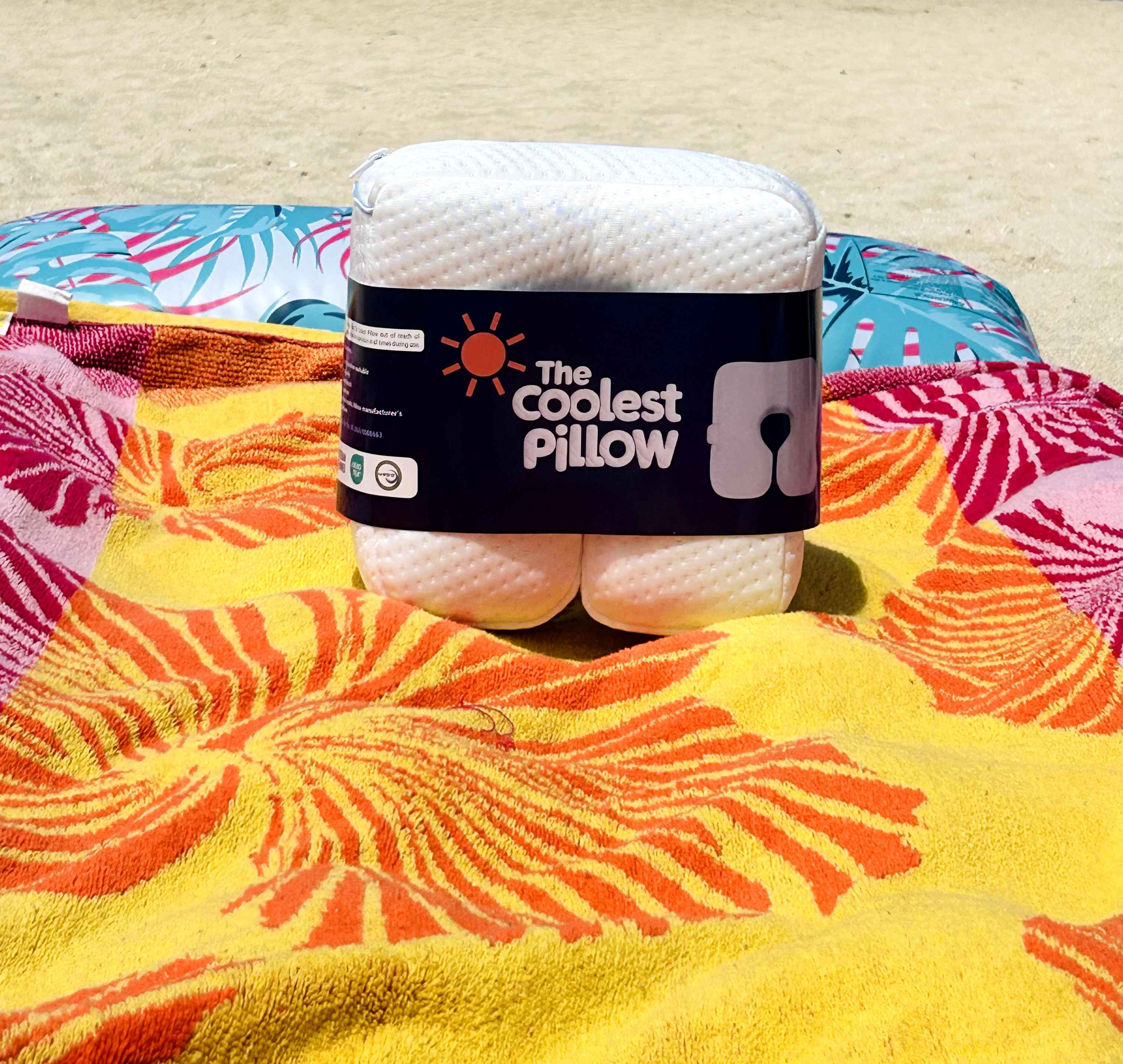 The Coolest Pillow, a cooling beach pillow, in branded packaging on a colorful floral towel, ideal for a refreshing beach rest and tan, by Bunzzup.