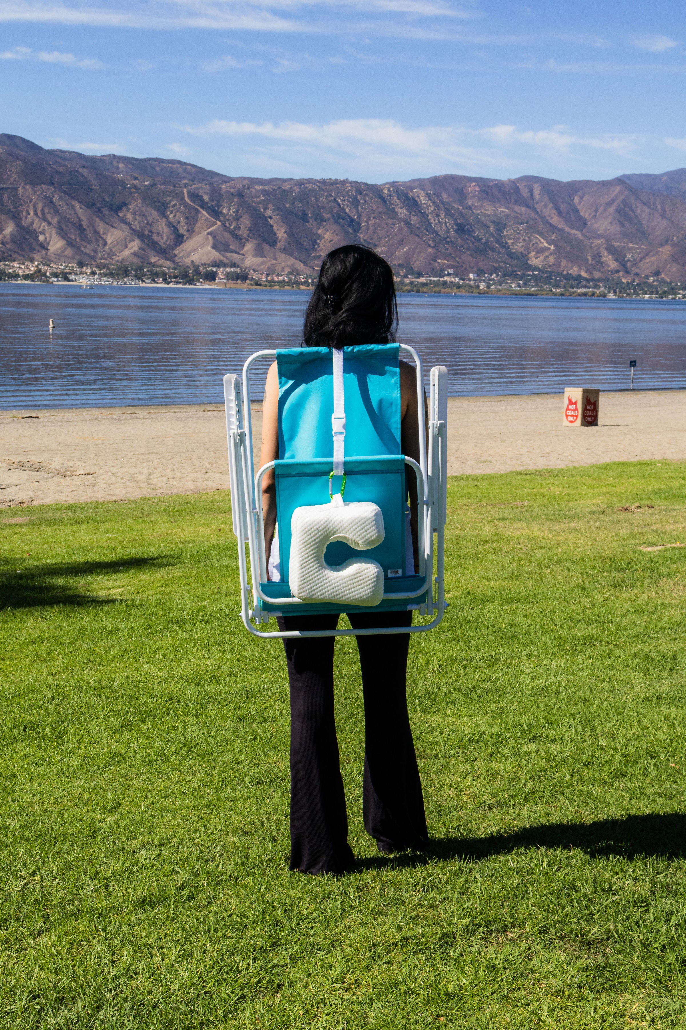 A person carries a blue beach chair with The Coolest Pillow, a cooling beach pillow with U shaped designed and memory foam, clipped with carabiner by a serene lake with mountains for comfy outdoor rest.