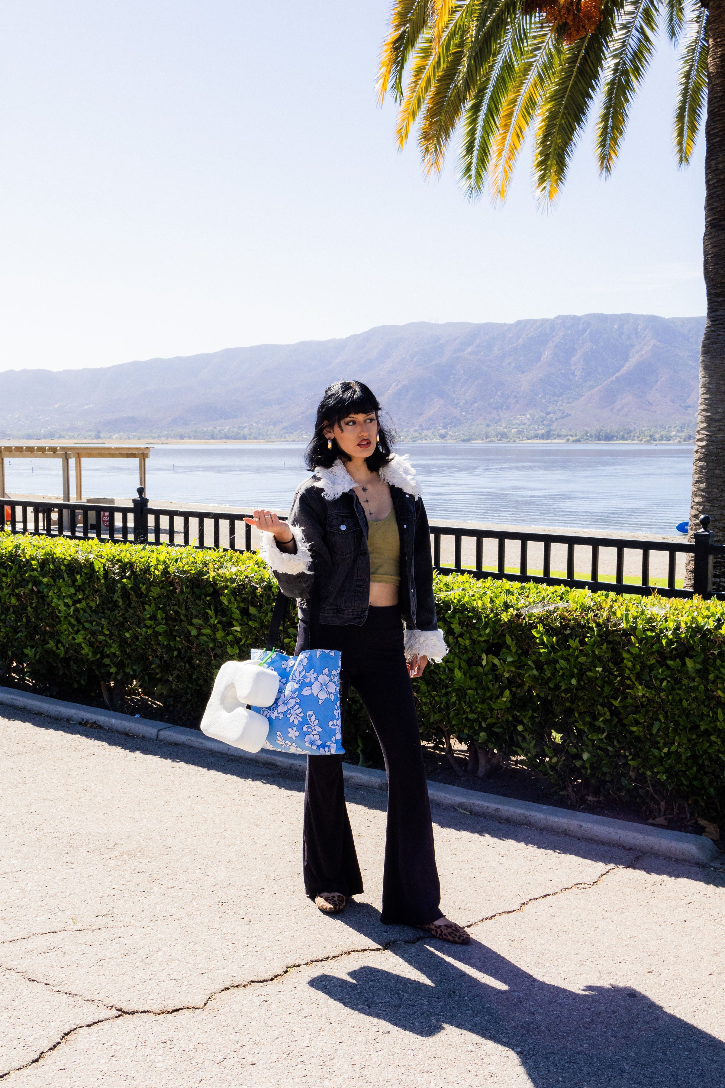 A stylish person in a fringed jacket with The Coolest Pillow, a cooling beach pillow, clipped to a blue tote bag by a serene lake with mountains.