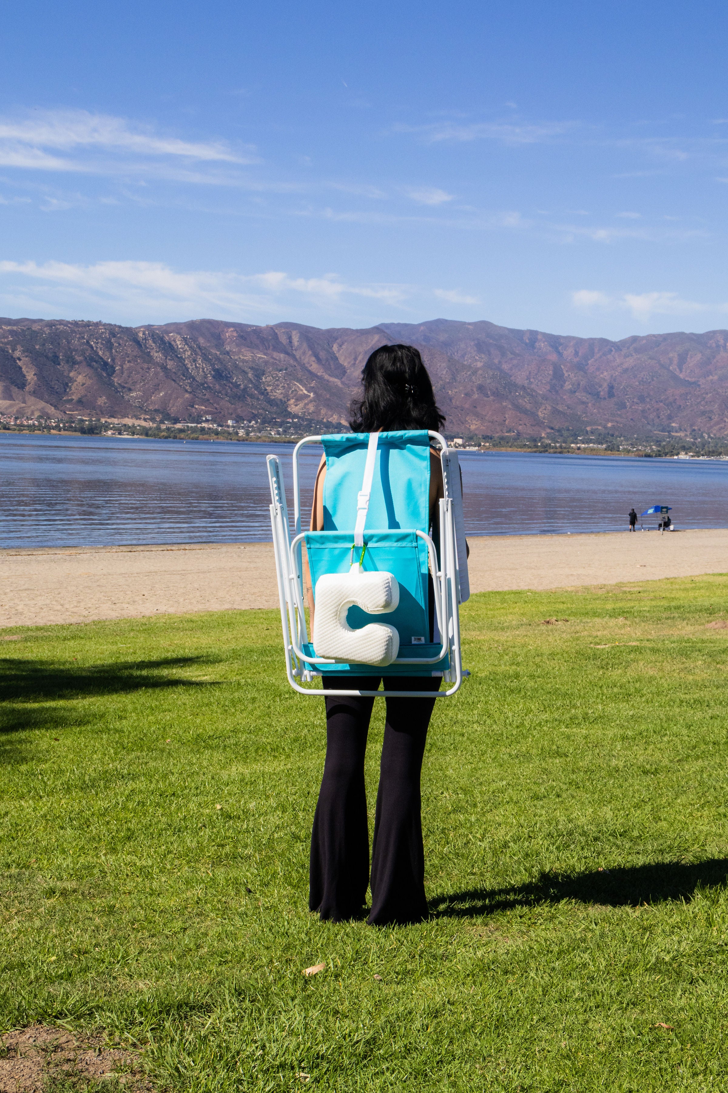 A person carries a blue beach chair with The Coolest Pillow, a cooling beach pillow with U shaped designed and memory foam, clipped with carabiner by a serene lake with mountains for comfy outdoor rest.